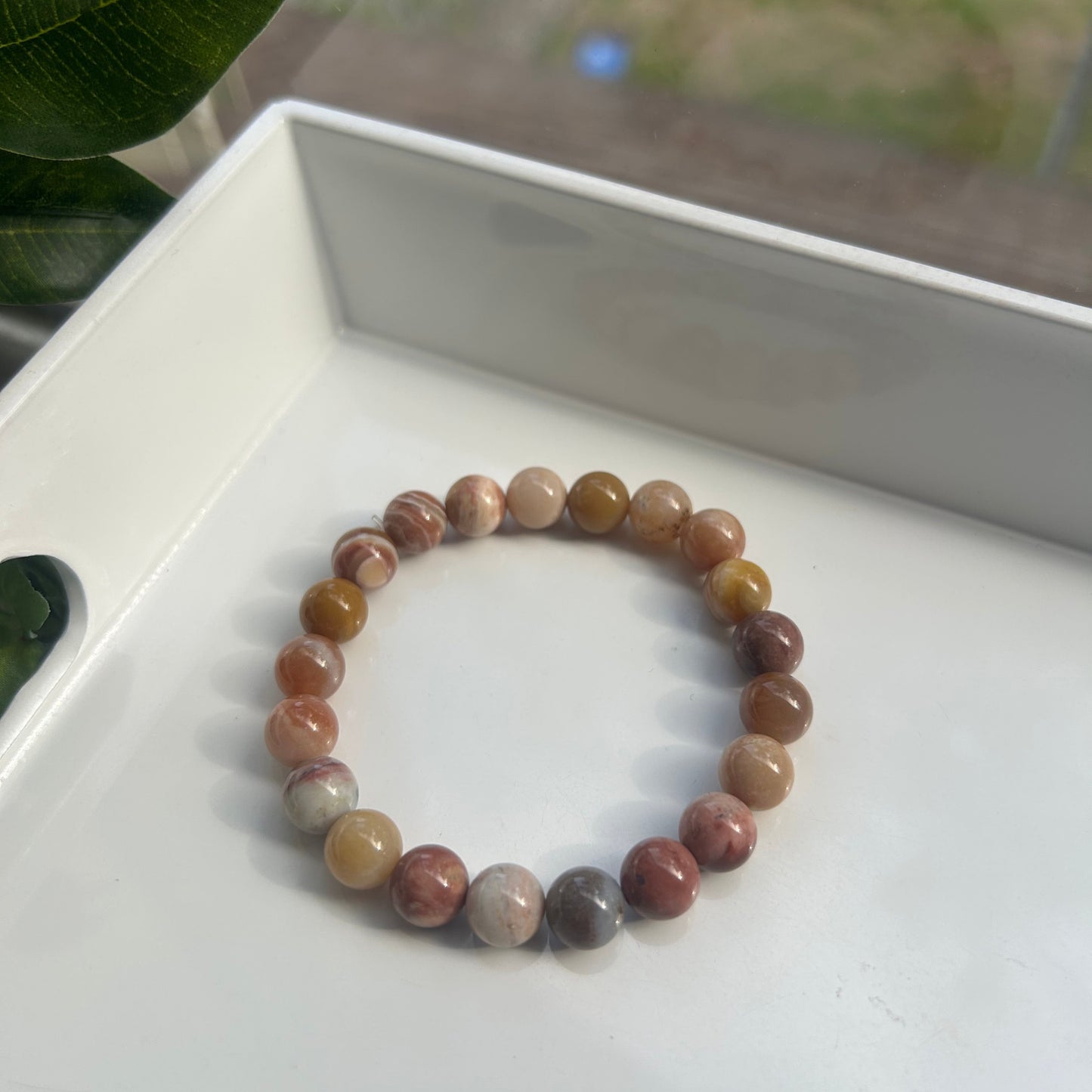 A beaded stretch bracelet made of polished round gemstone beads in earthy brown, beige, and muted pink tones, resting on a white tray.