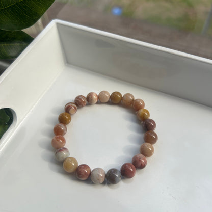 A beaded stretch bracelet made of polished round gemstone beads in earthy brown, beige, and muted pink tones, resting on a white tray.