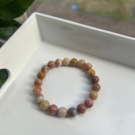 A beaded stretch bracelet made of polished round gemstone beads in earthy brown, beige, and muted pink tones, resting on a white tray.