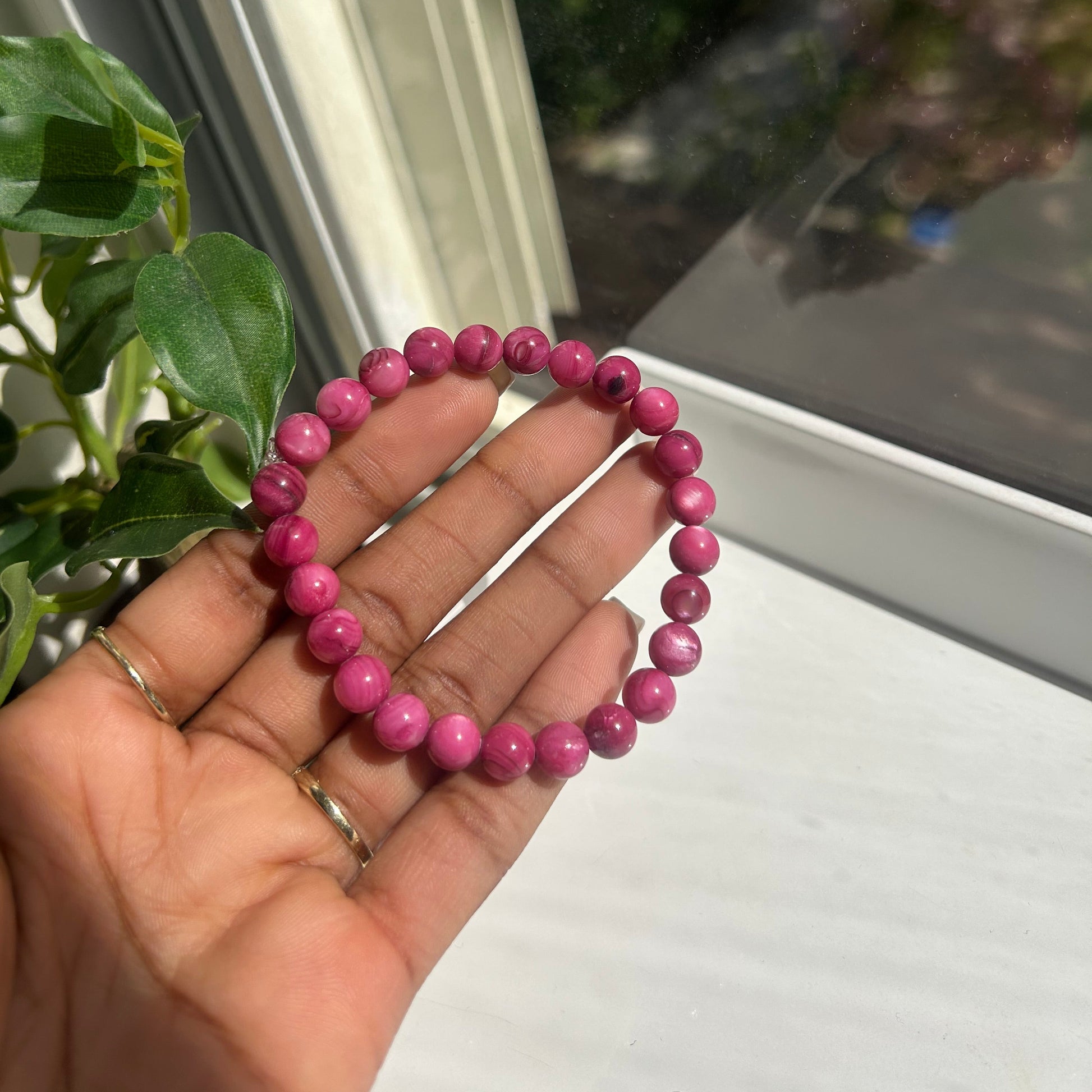 A pink beaded bracelet held in a person's hand near a window with green leaves in the foreground.