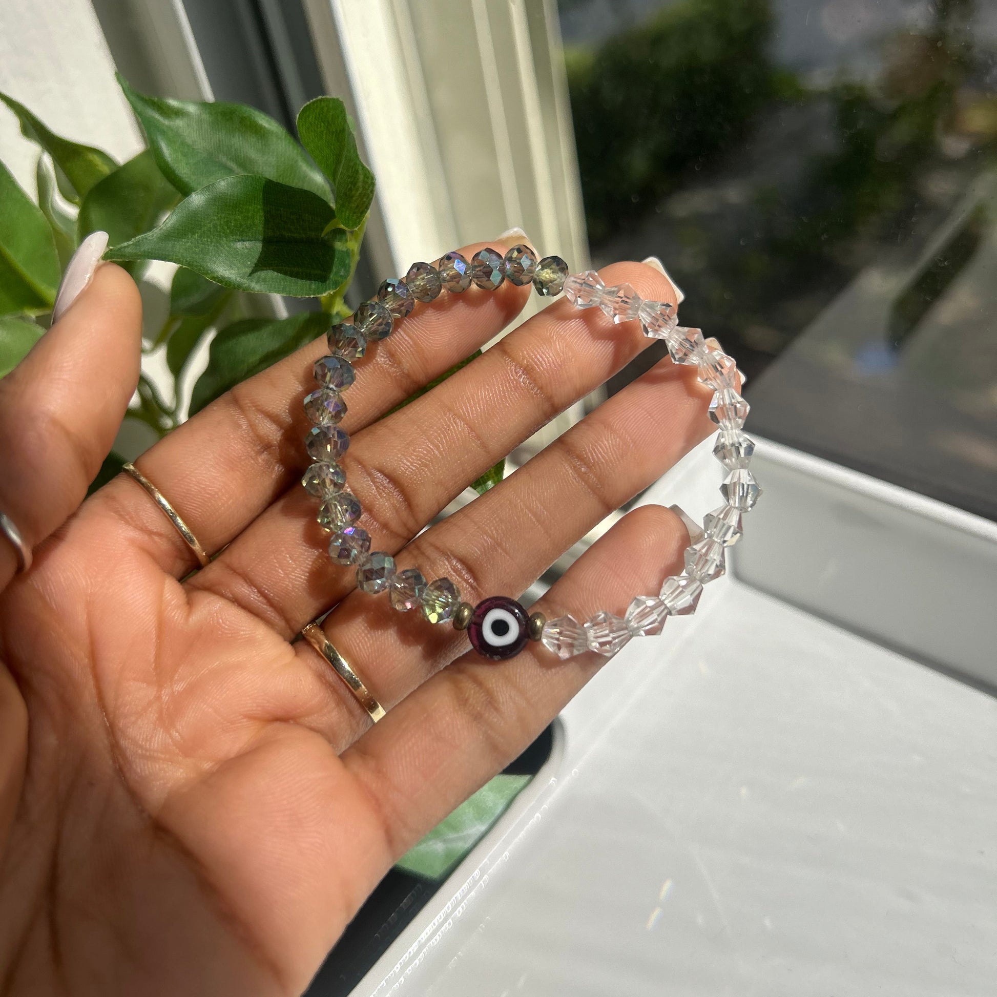 A person's hand holding a beaded stretch bracelet made of clear faceted beads with a small evil eye charm, set against a background of a window and green leaves.