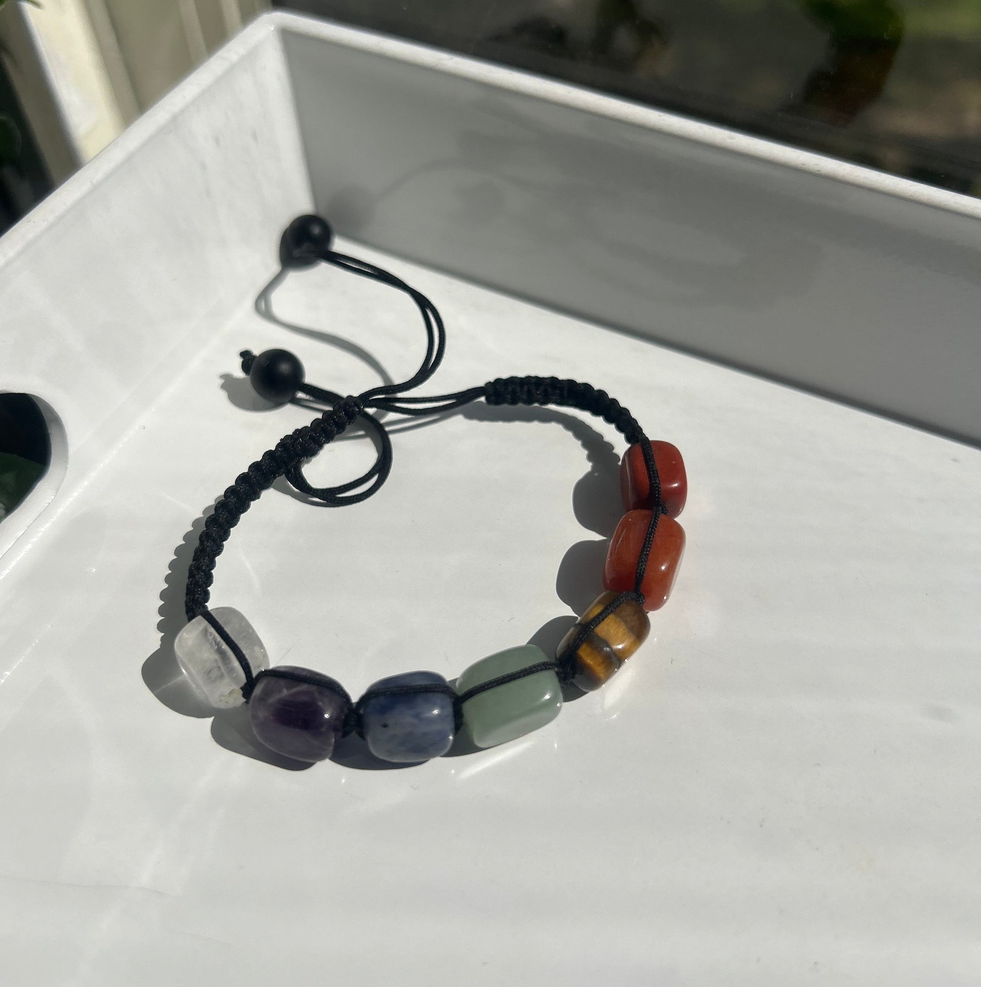 A beaded bracelet with a black braided cord and polished stone beads in orange, brown, green, purple, and clear, displayed on a white surface.