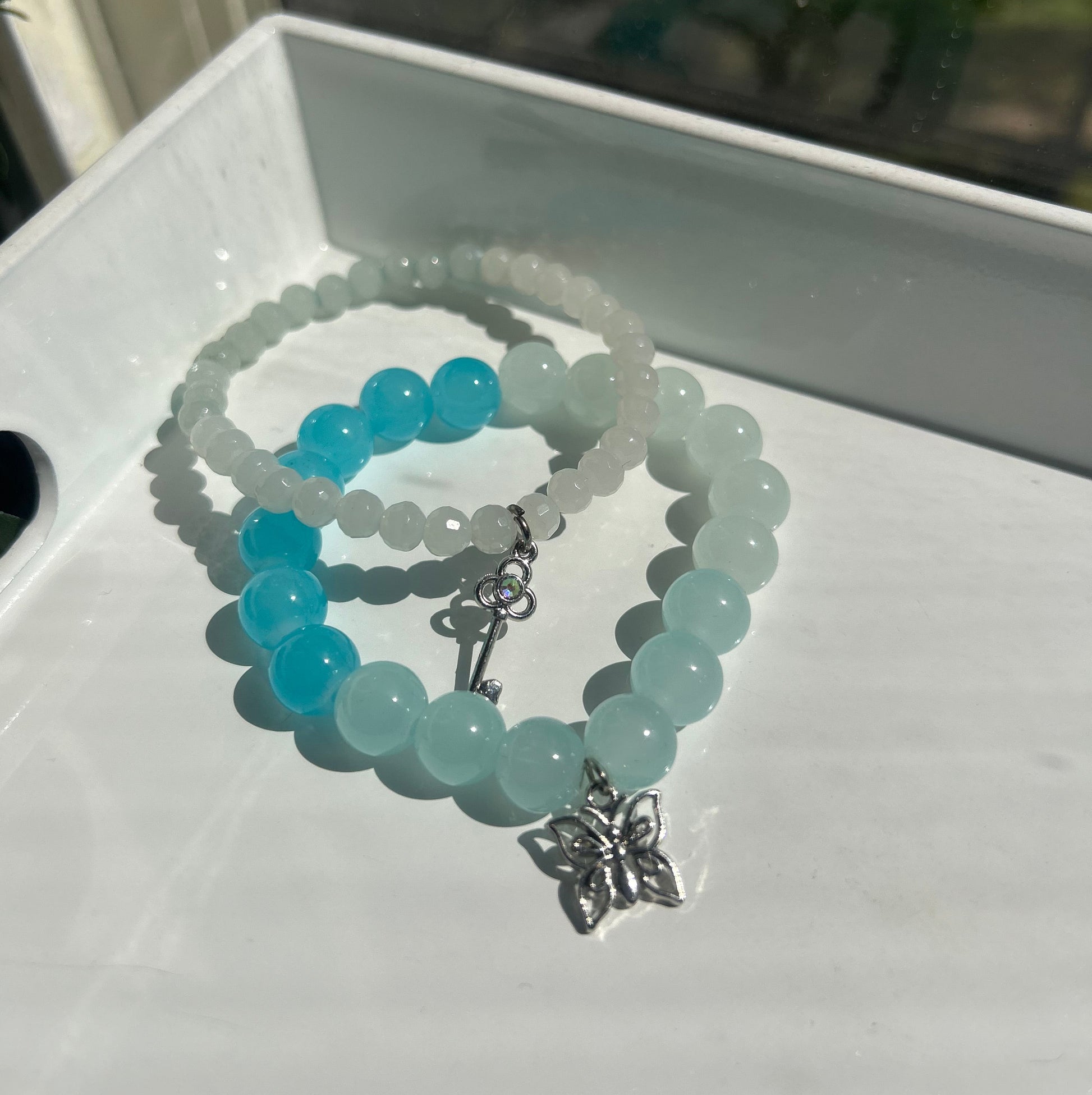 Two beaded bracelets displayed on a white tray. One bracelet features light blue-green round beads with a silver butterfly charm, and the other features translucent white beads with bright blue beads and a silver clasp.