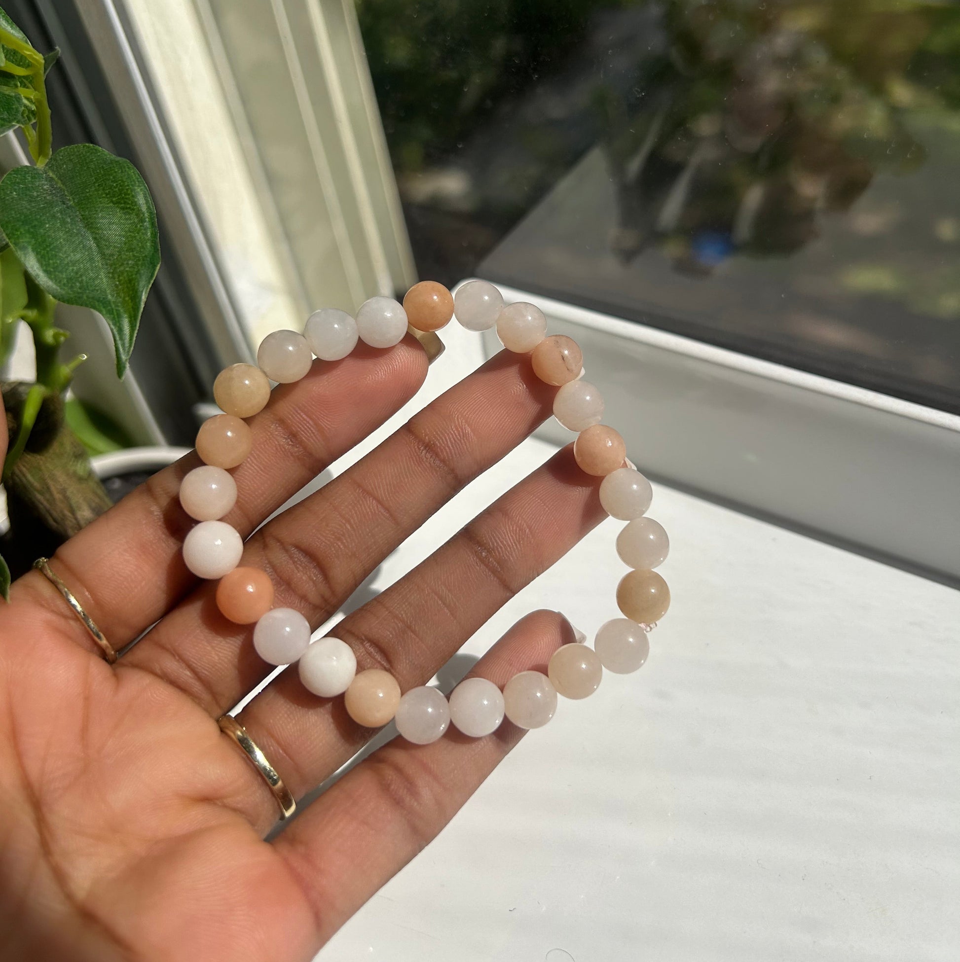 A beaded stretch bracelet made of smooth, round translucent beads in soft peach, cream, and white tones, held in a person's hand near a window.