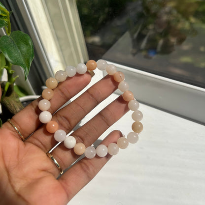 A beaded stretch bracelet made of smooth, round translucent beads in soft peach, cream, and white tones, held in a person's hand near a window.