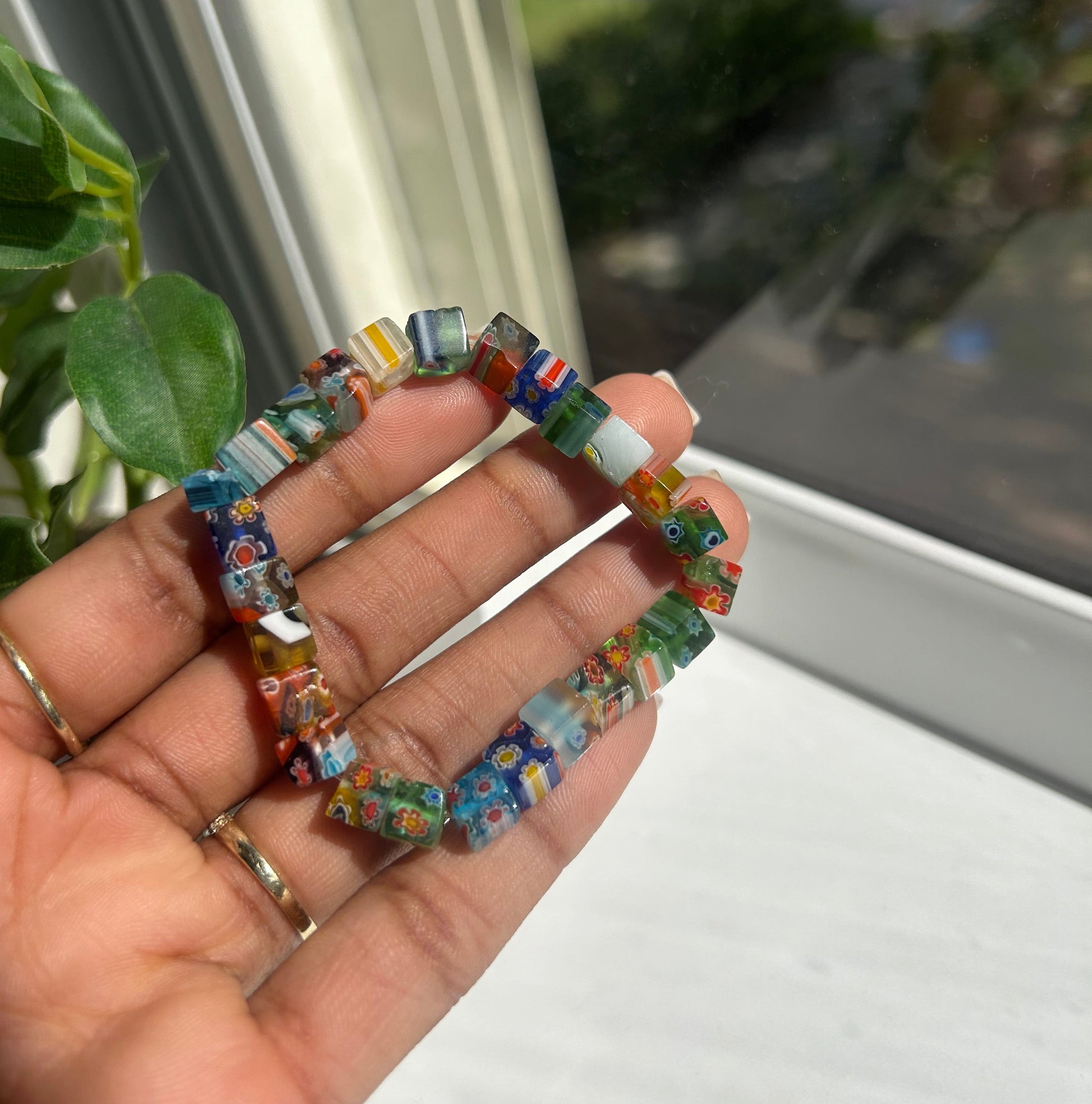 A hand holding a stretch bracelet made of small square glass beads with a colorful, marbled pattern. The background shows a window with green foliage outside.