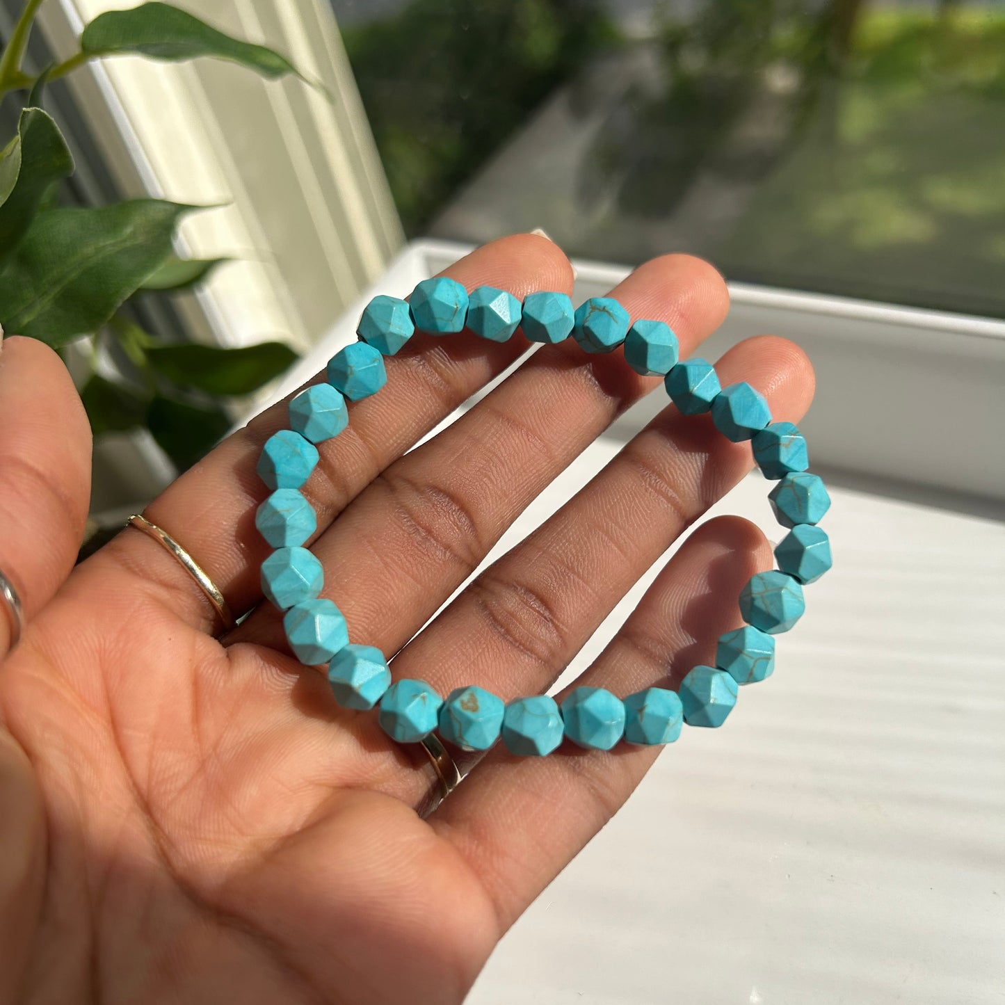 A beaded stretch bracelet made of faceted turquoise-blue beads, held in a person's hand near a window.
