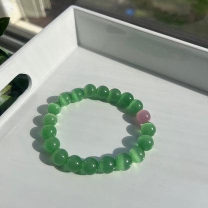 A beaded bracelet made of translucent light green round beads with one small pink accent bead, resting on a white surface.