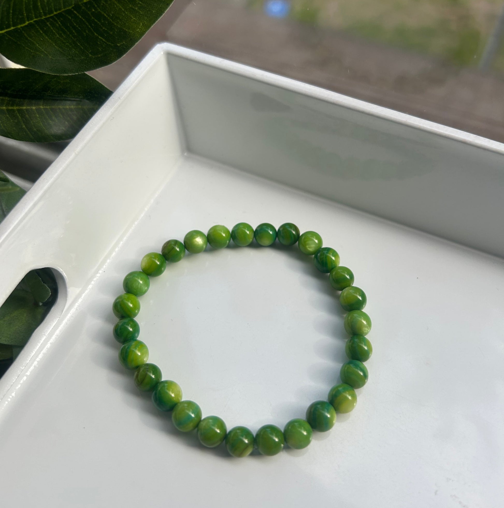 A green beaded bracelet made of polished round stones, resting inside a white tray with a blurred window and plant leaves in the background.