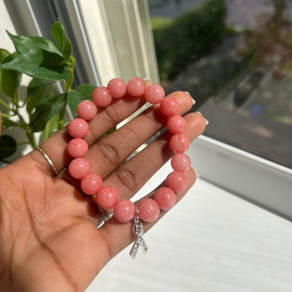 A person's hand holding a pink beaded bracelet with a small silver butterfly charm, set against a background of a window and green leaves.