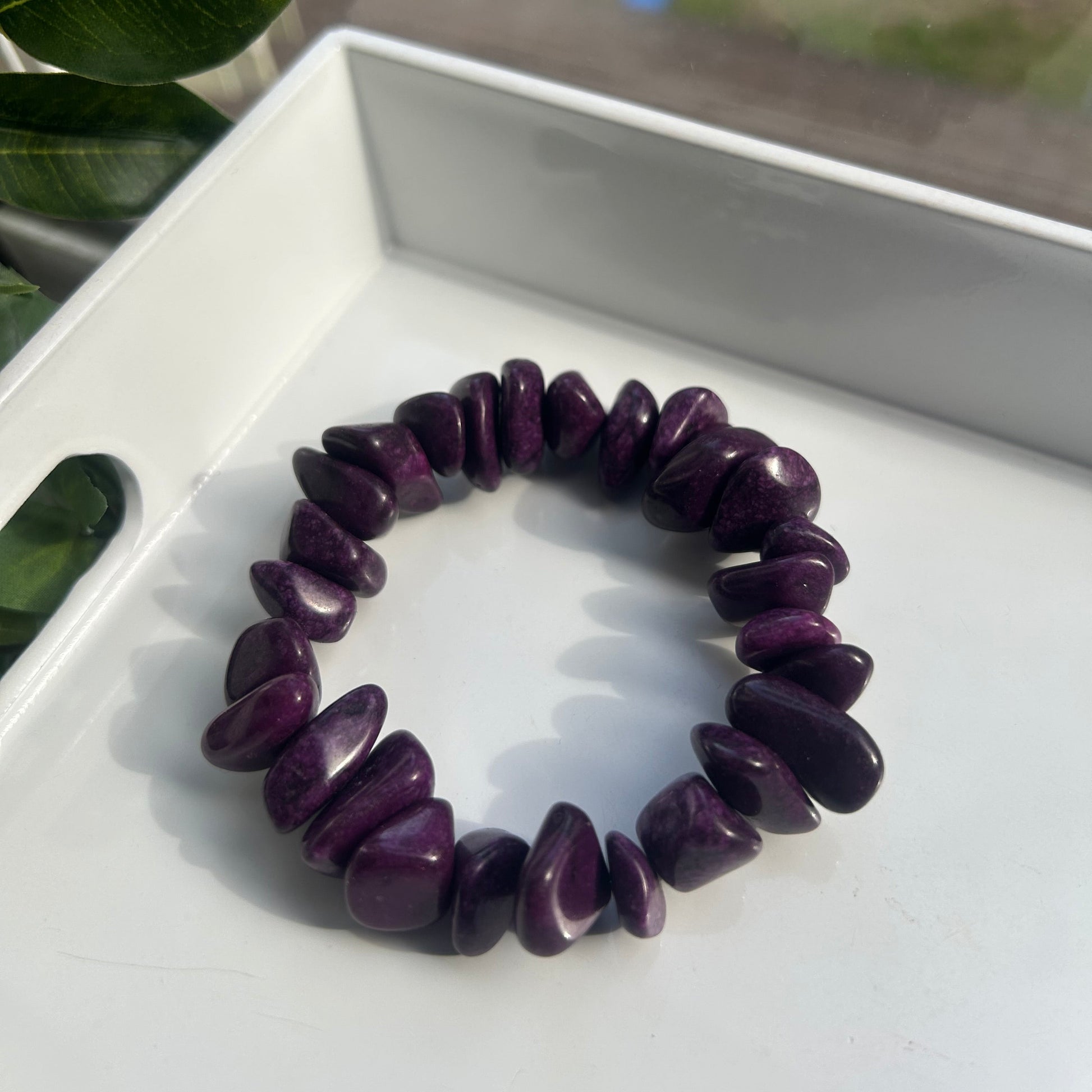 A dark purple tumbled stone chip bracelet displayed on a white tray, with a blurred window and green leaves in the background.