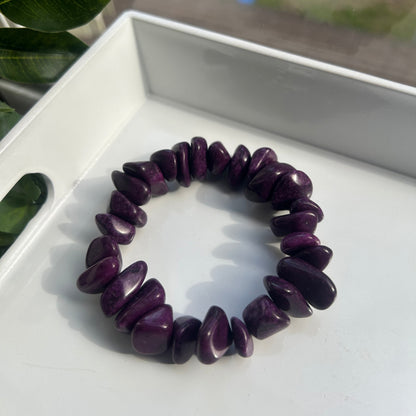 A dark purple tumbled stone chip bracelet displayed on a white tray, with a blurred window and green leaves in the background.