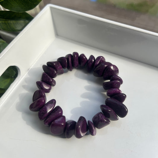 A dark purple tumbled stone chip bracelet displayed on a white tray, with a blurred window and green leaves in the background.