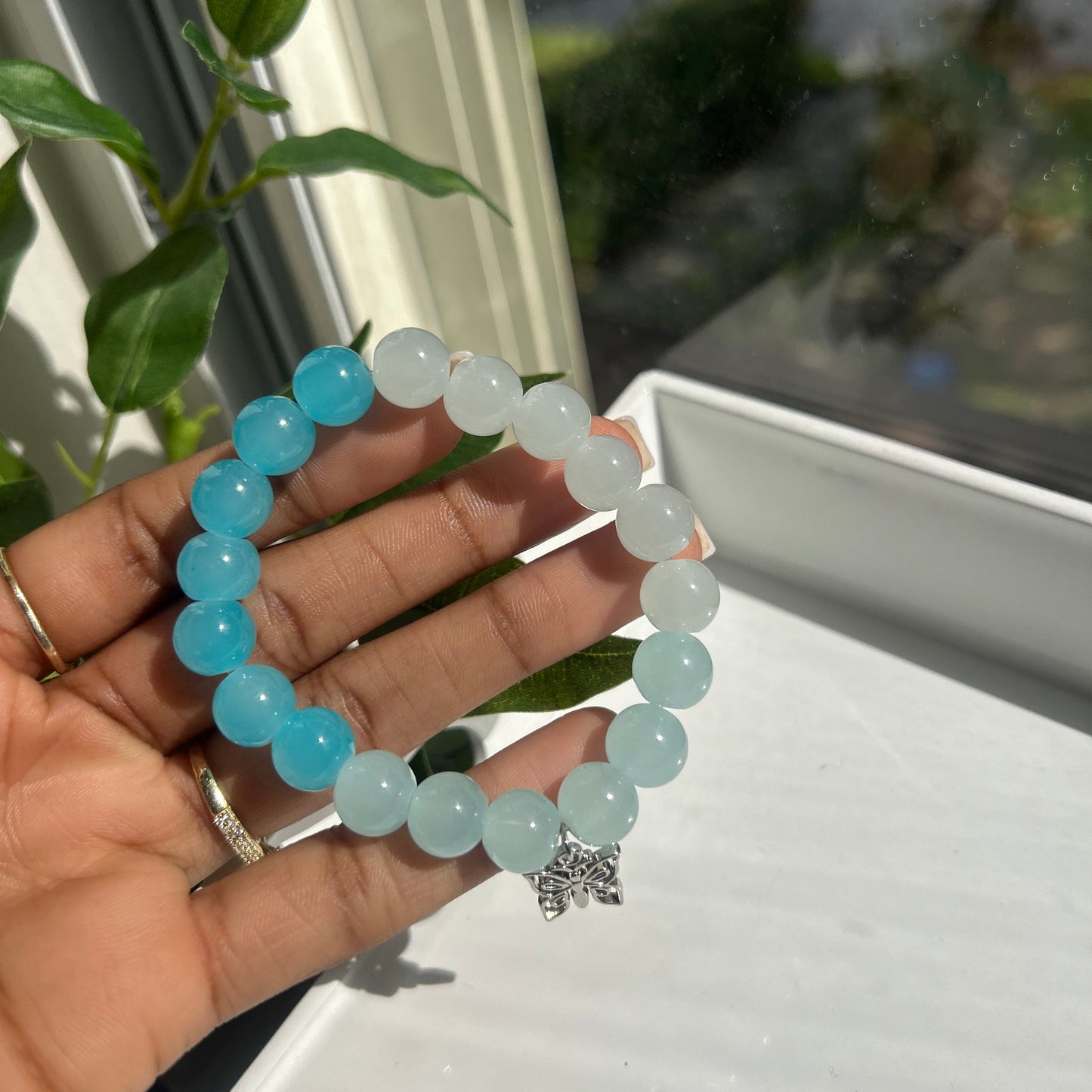 A beaded stretch bracelet held in a hand, featuring light blue and milky white round beads with a small silver-toned star charm, set against a bright window background.