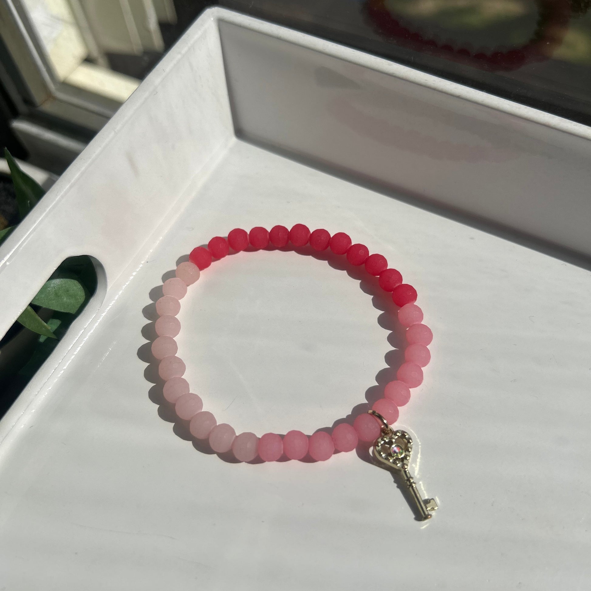 A beaded stretch bracelet with a gradient of pink and red beads, featuring a silver key-shaped clasp, displayed on a white surface.