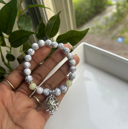 A hand holding a beaded stretch bracelet made of white and gray marbled beads, with a silver-toned lotus charm and a small pale green bead accent, shown near a window with green leaves.