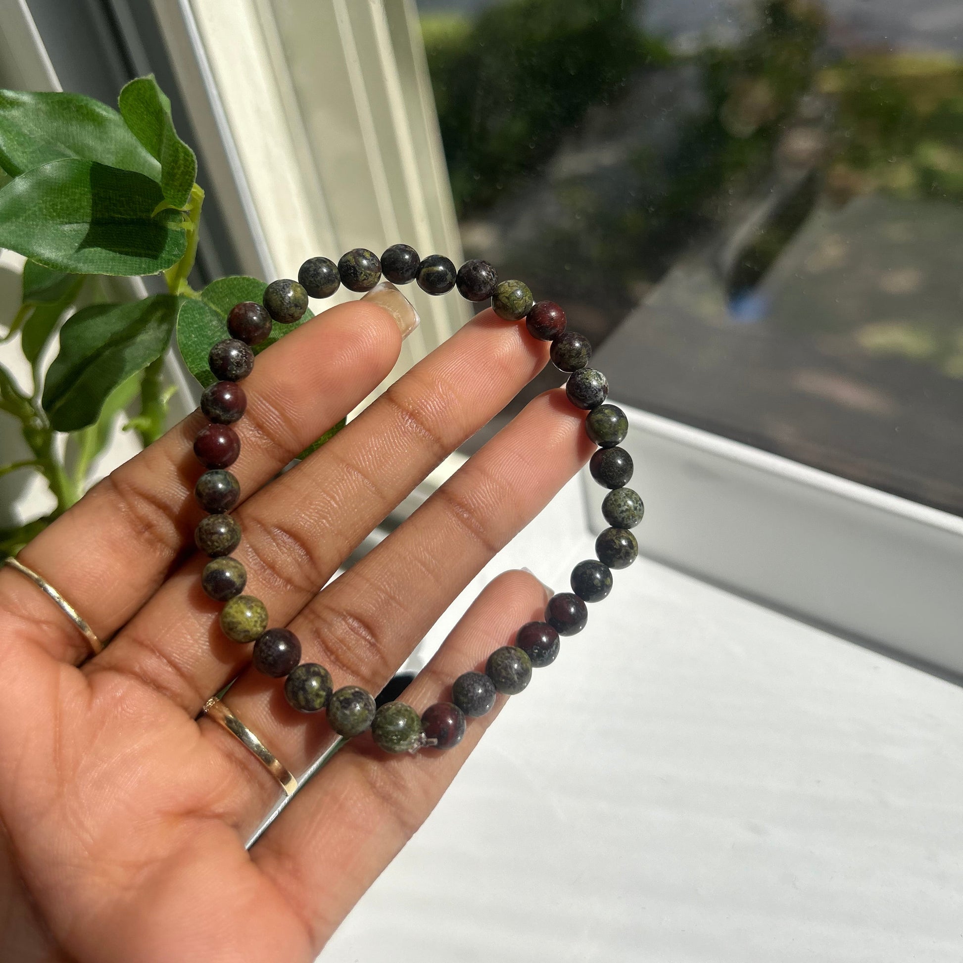 A beaded bracelet made of dark, polished round stones in shades of green, gray, and burgundy, held in a person's hand near a window.