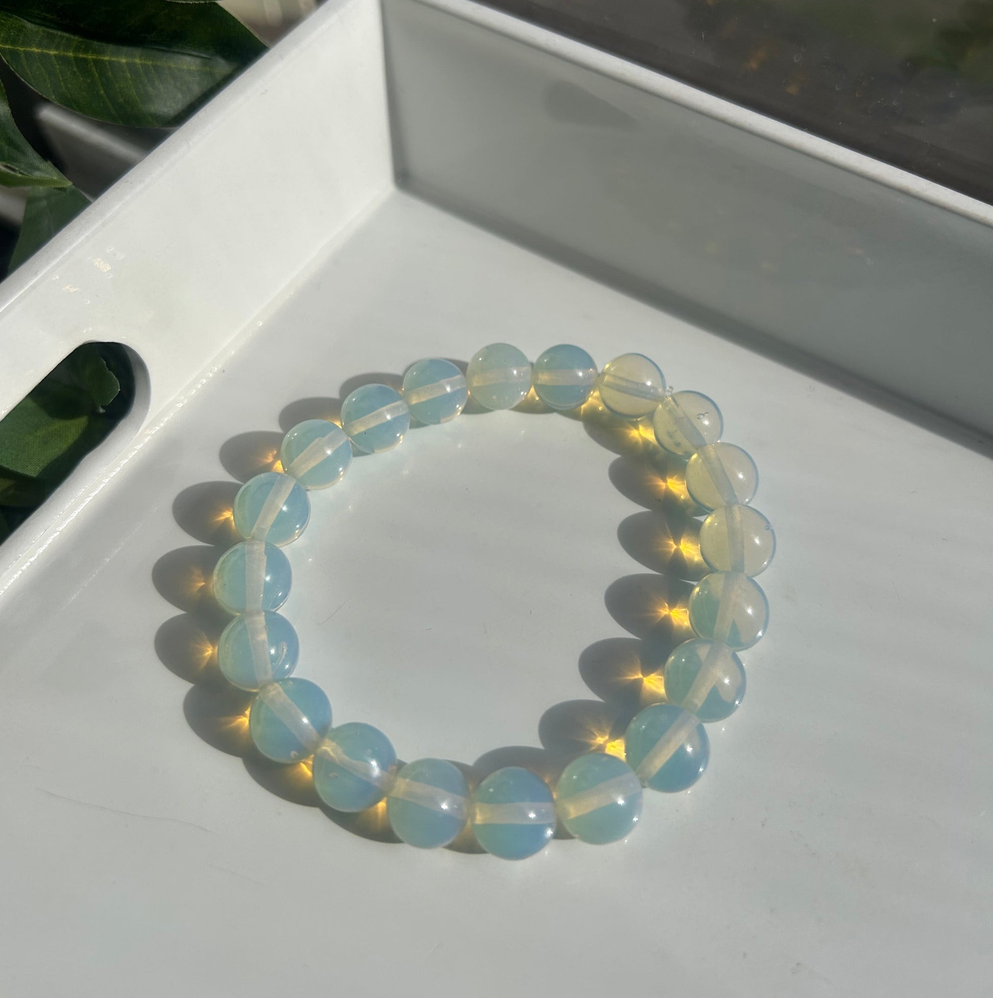 A beaded stretch bracelet made of smooth, translucent light blue and white oval beads, resting on a white tray with green leaves in the foreground.