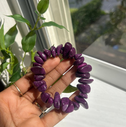 A purple tumbled stone beaded bracelet held in a person's hand near a window with green leaves in the foreground.