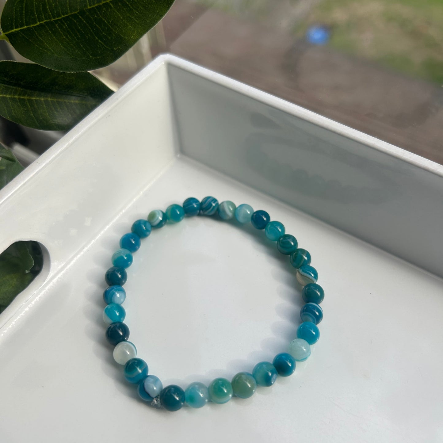 A beaded stretch bracelet made of round blue-green gemstone beads, resting on a white tray with green leaves in the foreground.