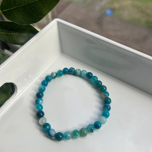 A beaded stretch bracelet made of round blue-green gemstone beads, resting on a white tray with green leaves in the foreground.