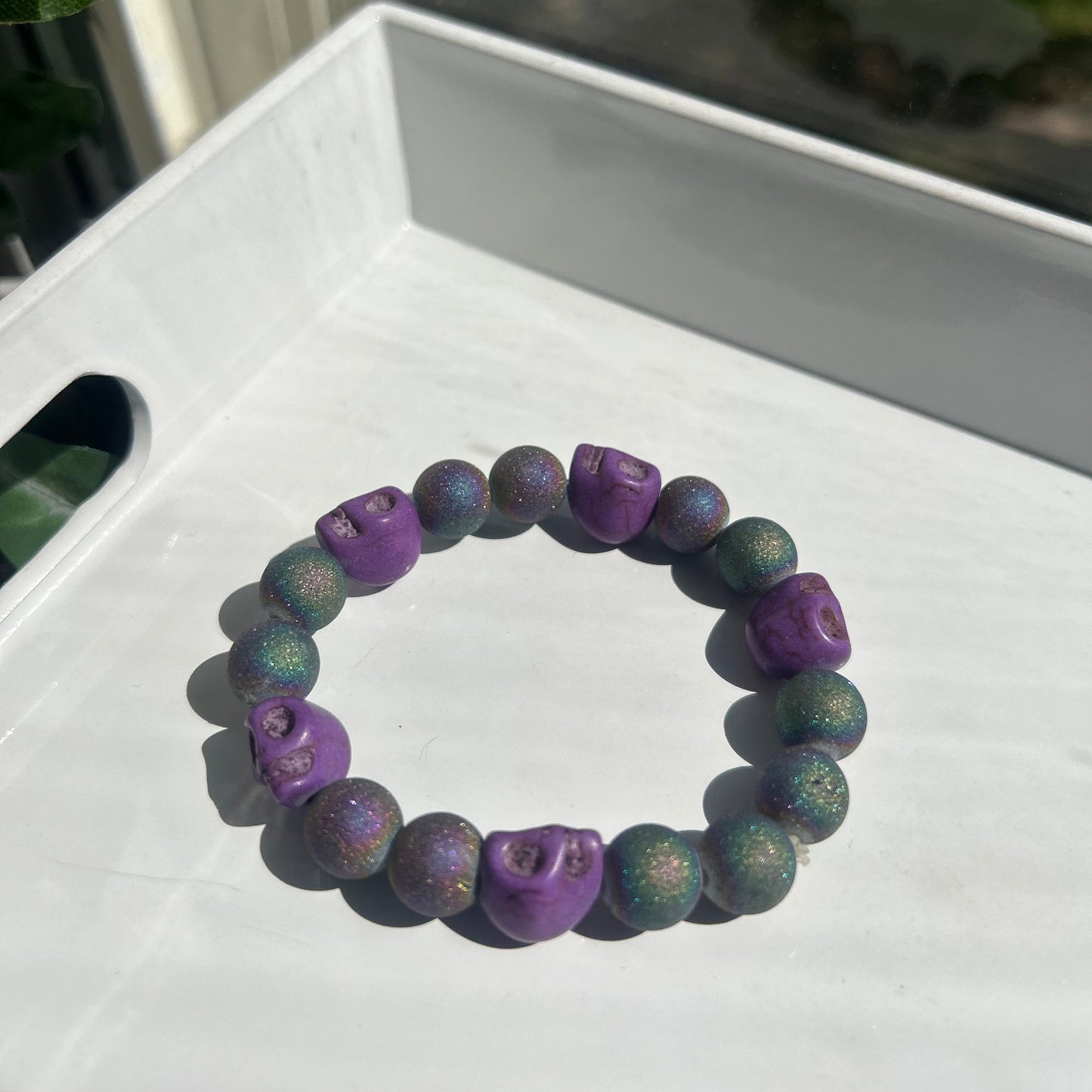 A beaded bracelet with alternating purple and green glittery round beads, featuring three purple skull-shaped beads, displayed on a white tray.