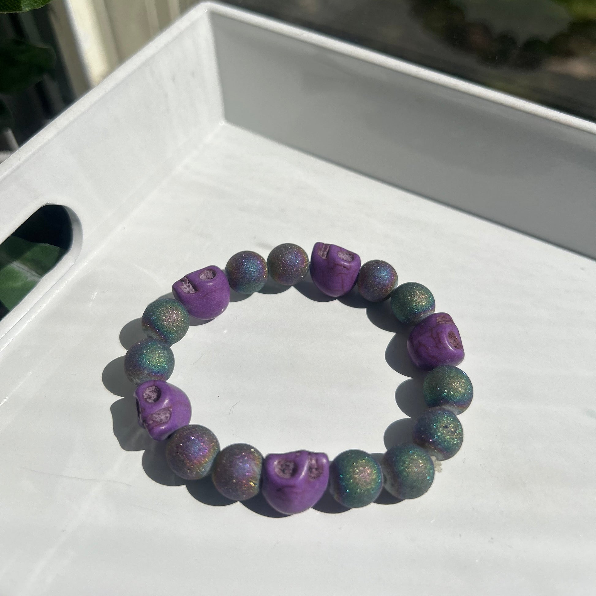 A beaded bracelet with alternating purple and green glittery round beads, featuring three purple skull-shaped beads, displayed on a white tray.