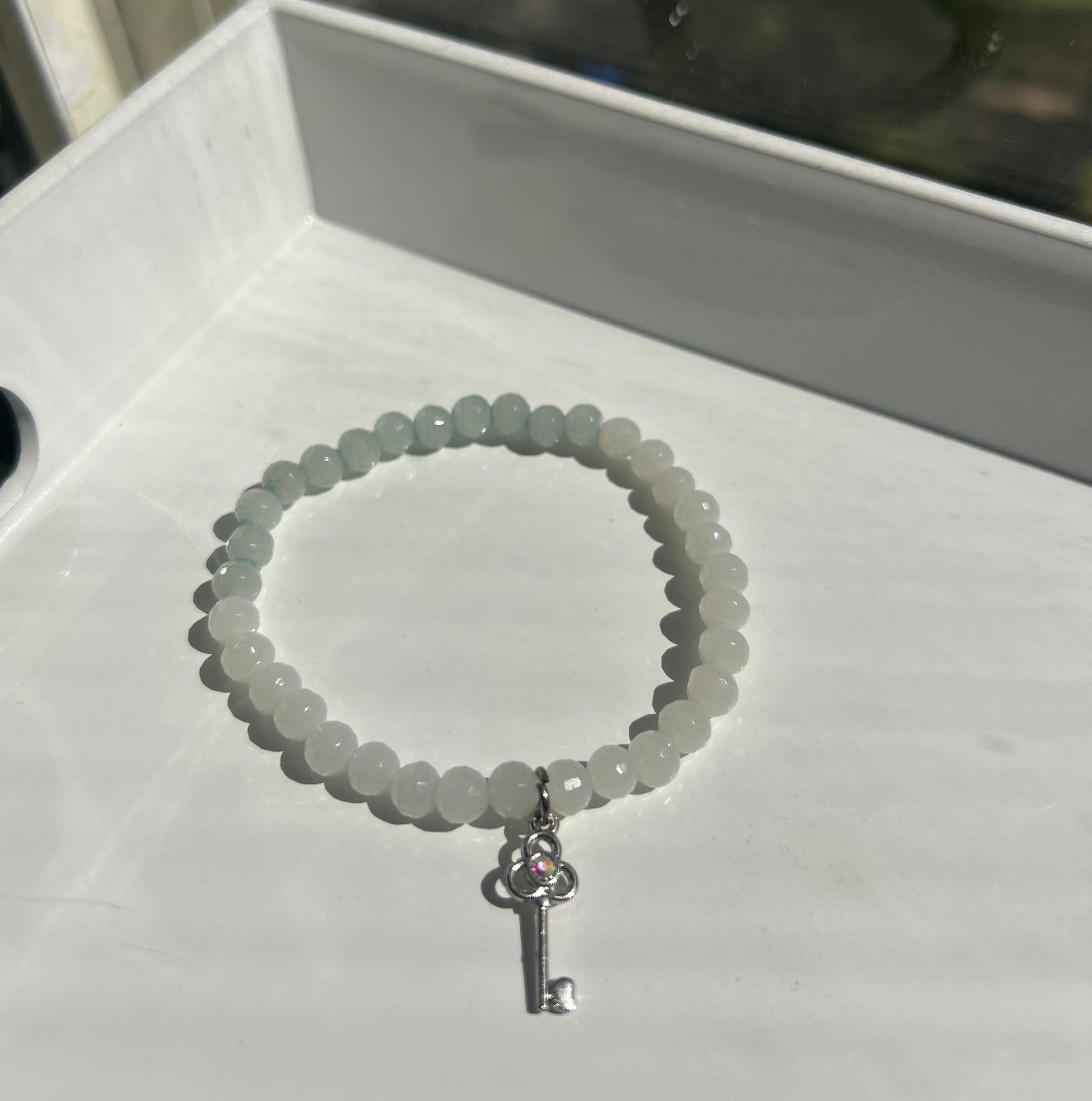 A beaded bracelet made of small pale gray-green round beads with a silver key-shaped clasp, displayed on a white surface.