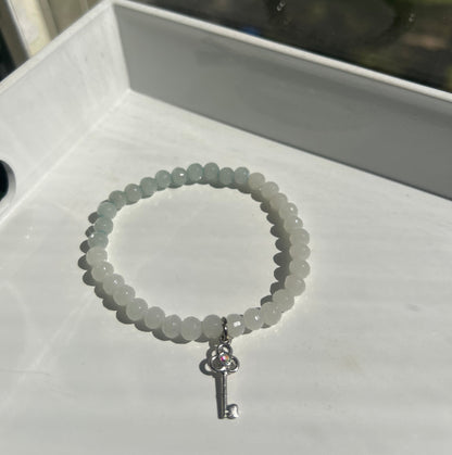 A beaded bracelet made of small pale gray-green round beads with a silver key-shaped clasp, displayed on a white surface.