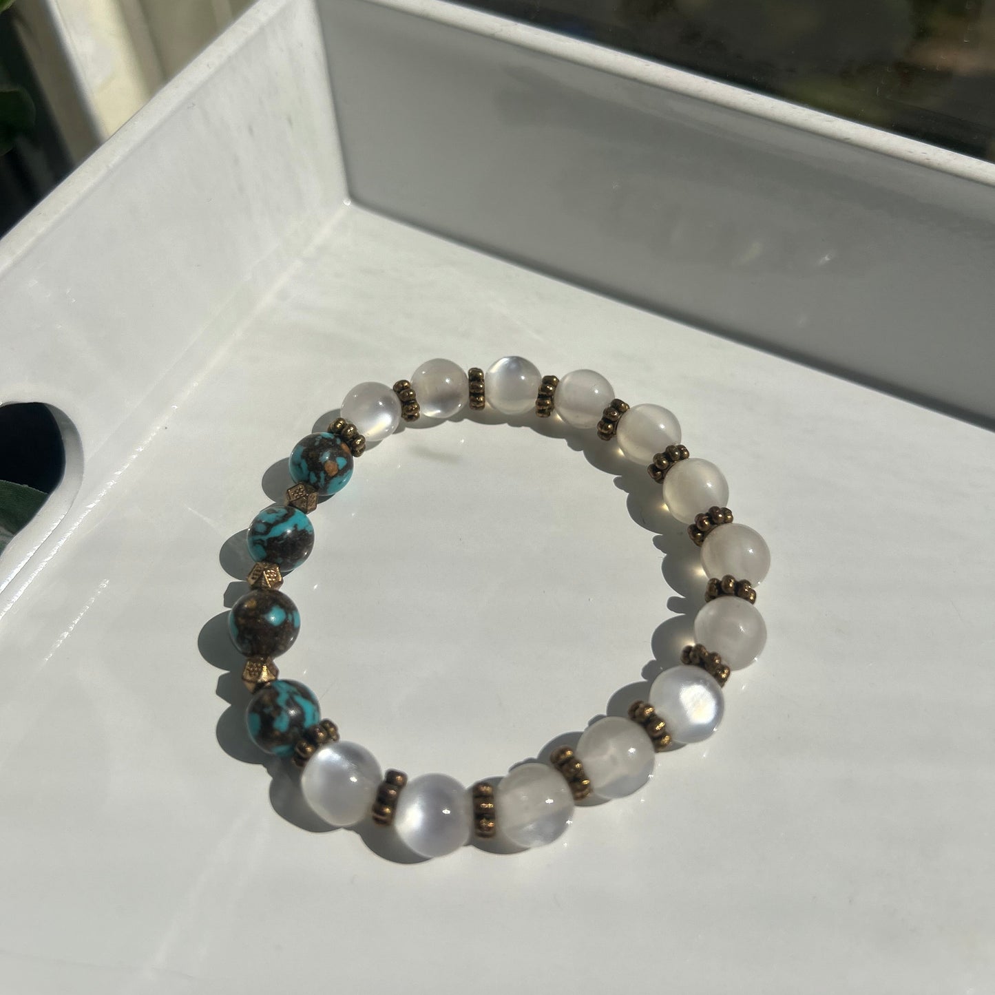 A beaded stretch bracelet featuring white moonstone beads, dark brown metallic spacer beads, and two turquoise-blue beads with bronze accents, displayed on a white tray.