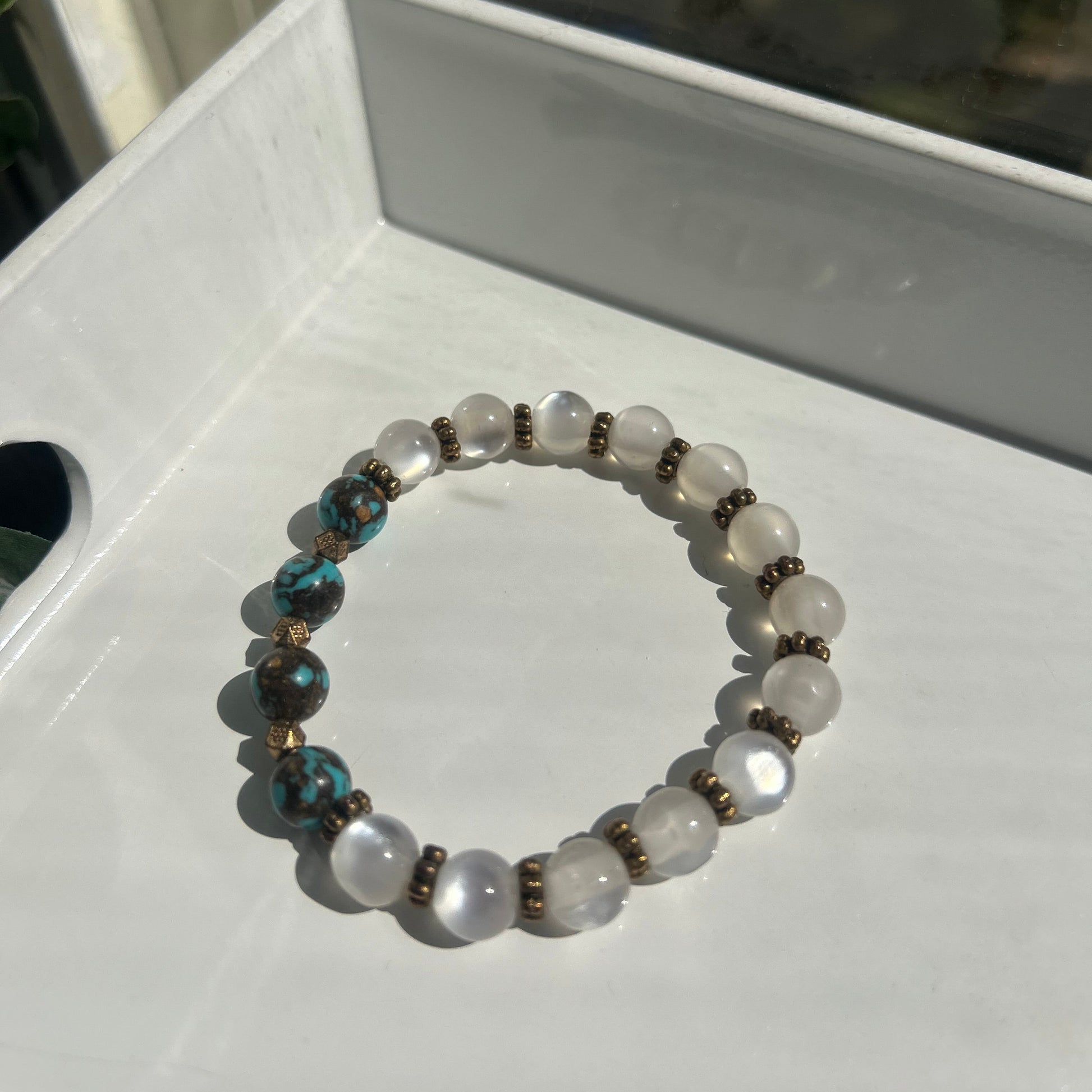 A beaded stretch bracelet featuring white moonstone beads, dark brown metallic spacer beads, and two turquoise-blue beads with bronze accents, displayed on a white tray.