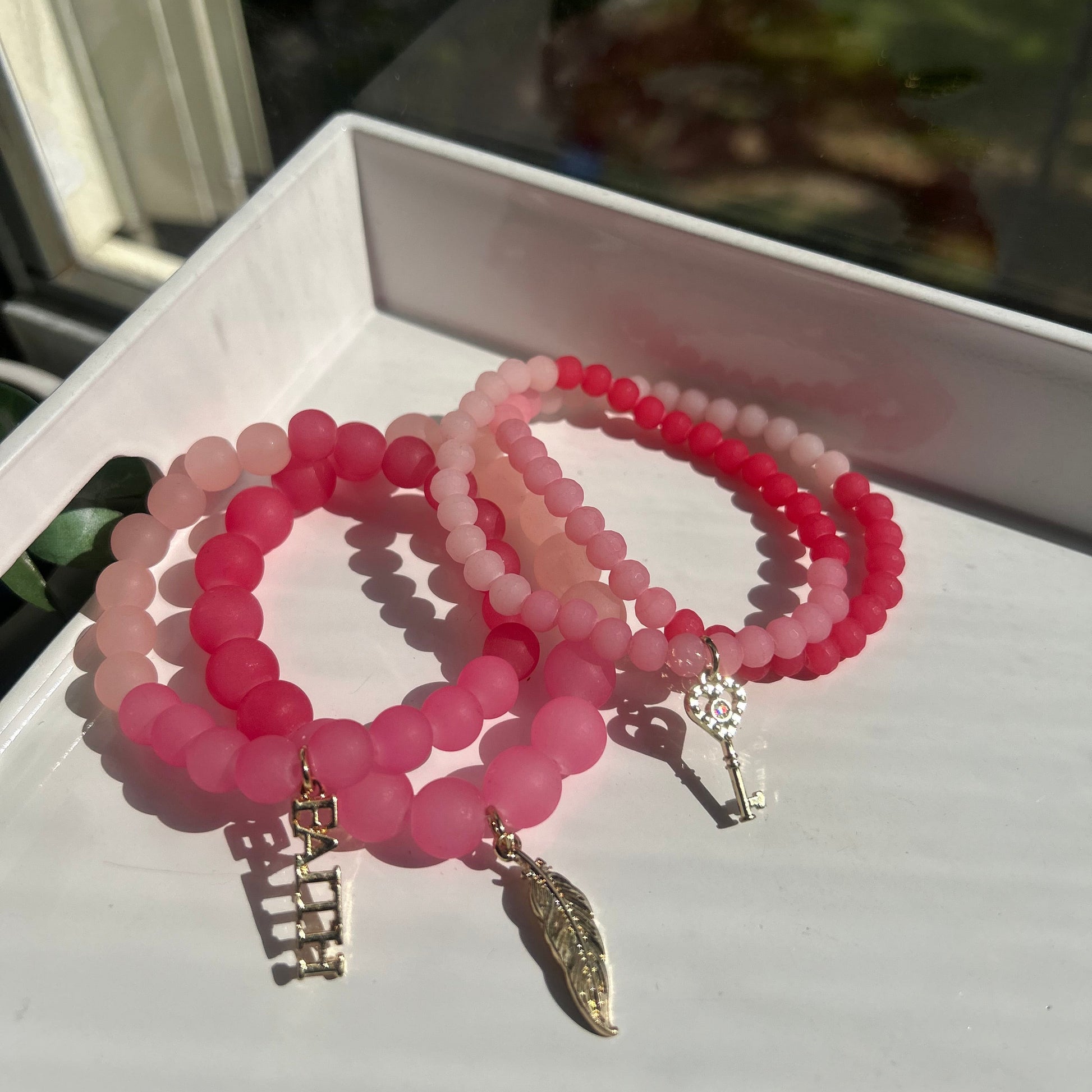 Three beaded bracelets in shades of pink and red, featuring gold-toned charms including a feather and the word 'BATCH', displayed on a white surface.