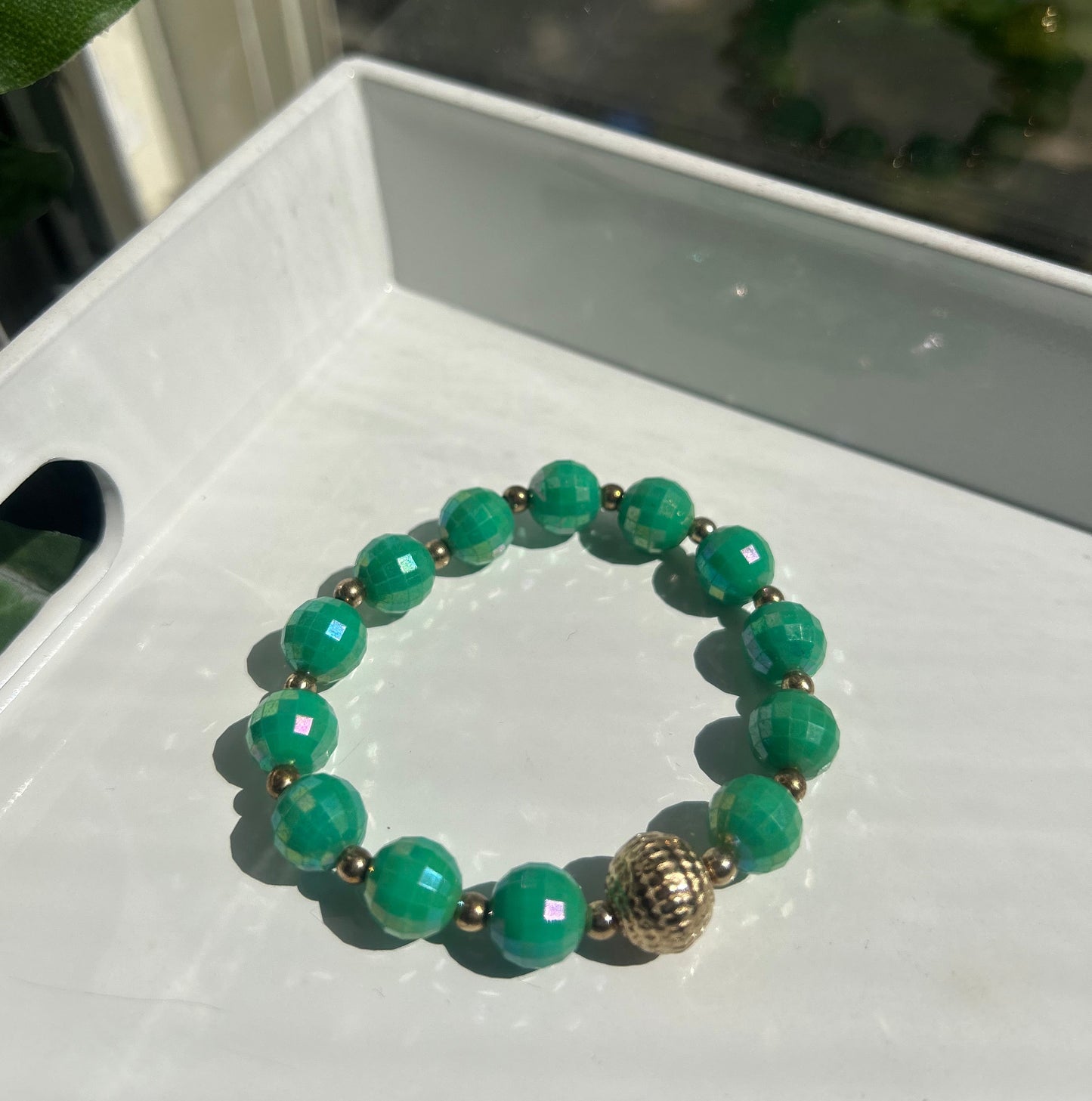 A beaded stretch bracelet made of green faceted beads with small metallic spacer beads and a textured gold-toned accent bead, resting on a white surface.