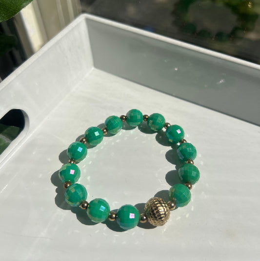 A beaded stretch bracelet made of green faceted beads with small metallic spacer beads and a textured gold-toned accent bead, resting on a white surface.