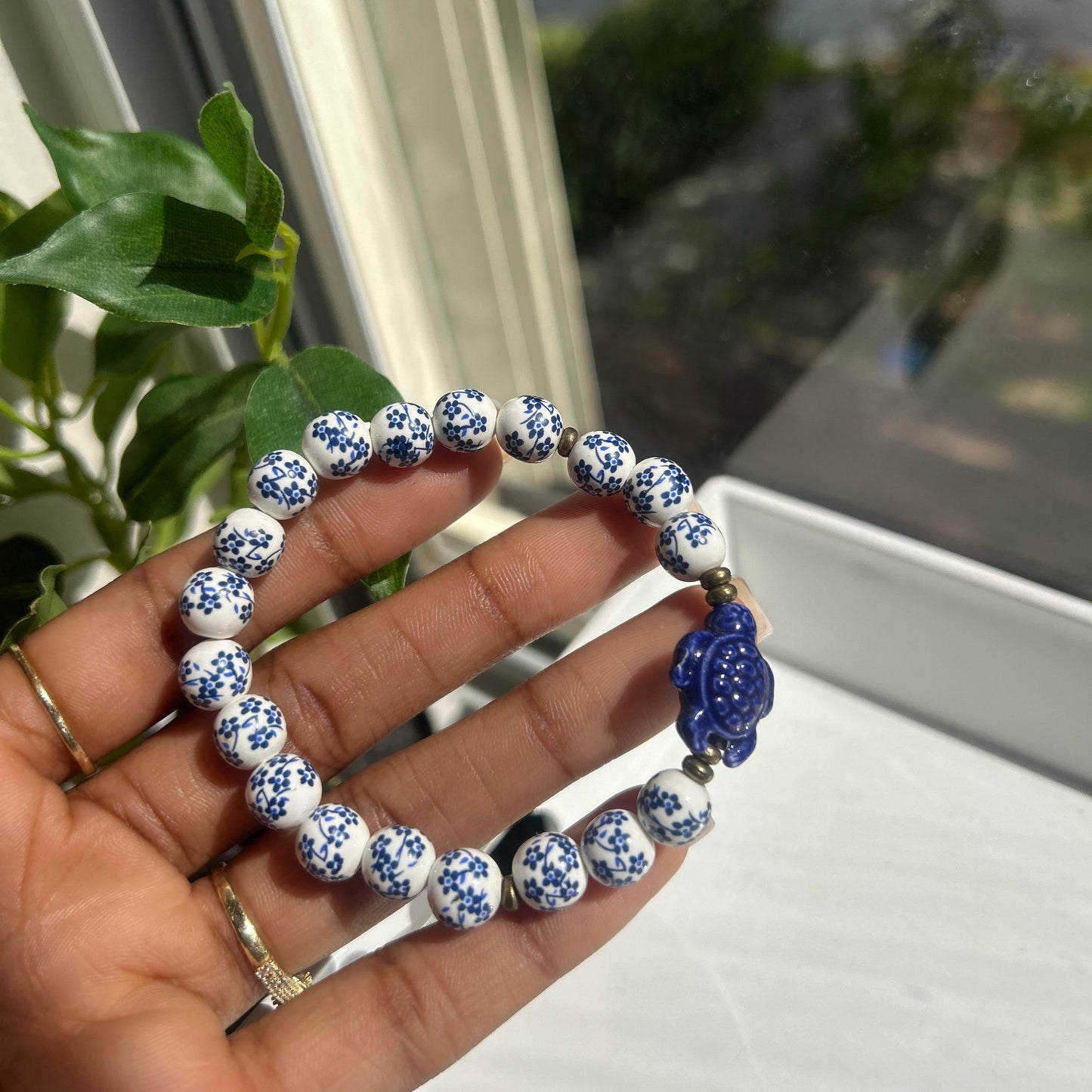 A hand holding a beaded bracelet with white and blue patterned round beads, a blue flower-shaped charm, and small gold spacer beads, photographed near a window with green leaves in the background.