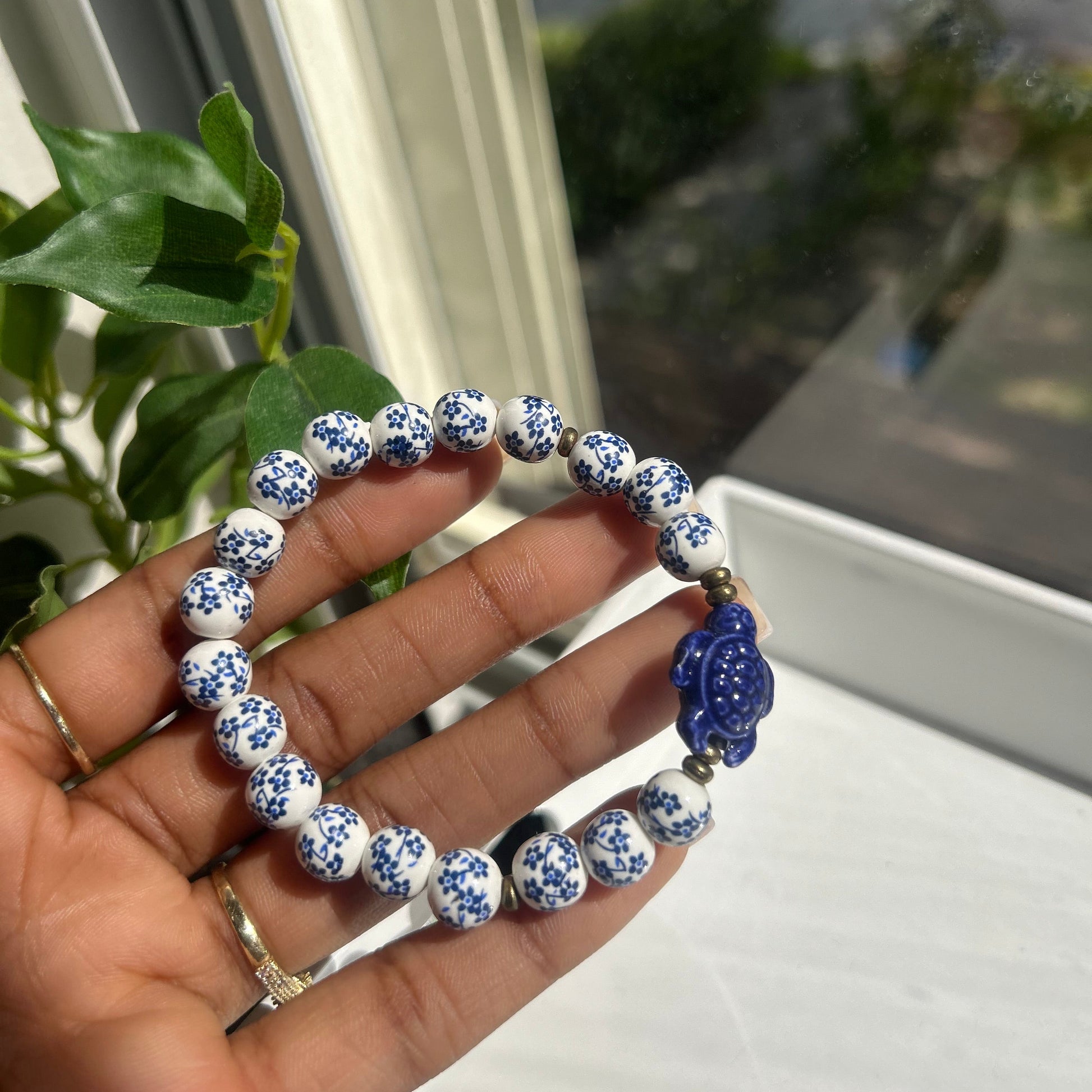 A hand holding a beaded bracelet with white and blue patterned round beads, a blue flower-shaped charm, and small gold spacer beads, photographed near a window with green leaves in the background.