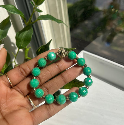 A beaded bracelet with round turquoise-green beads and a small metallic silver accent, held in a person's hand near a window with green leaves in the foreground.