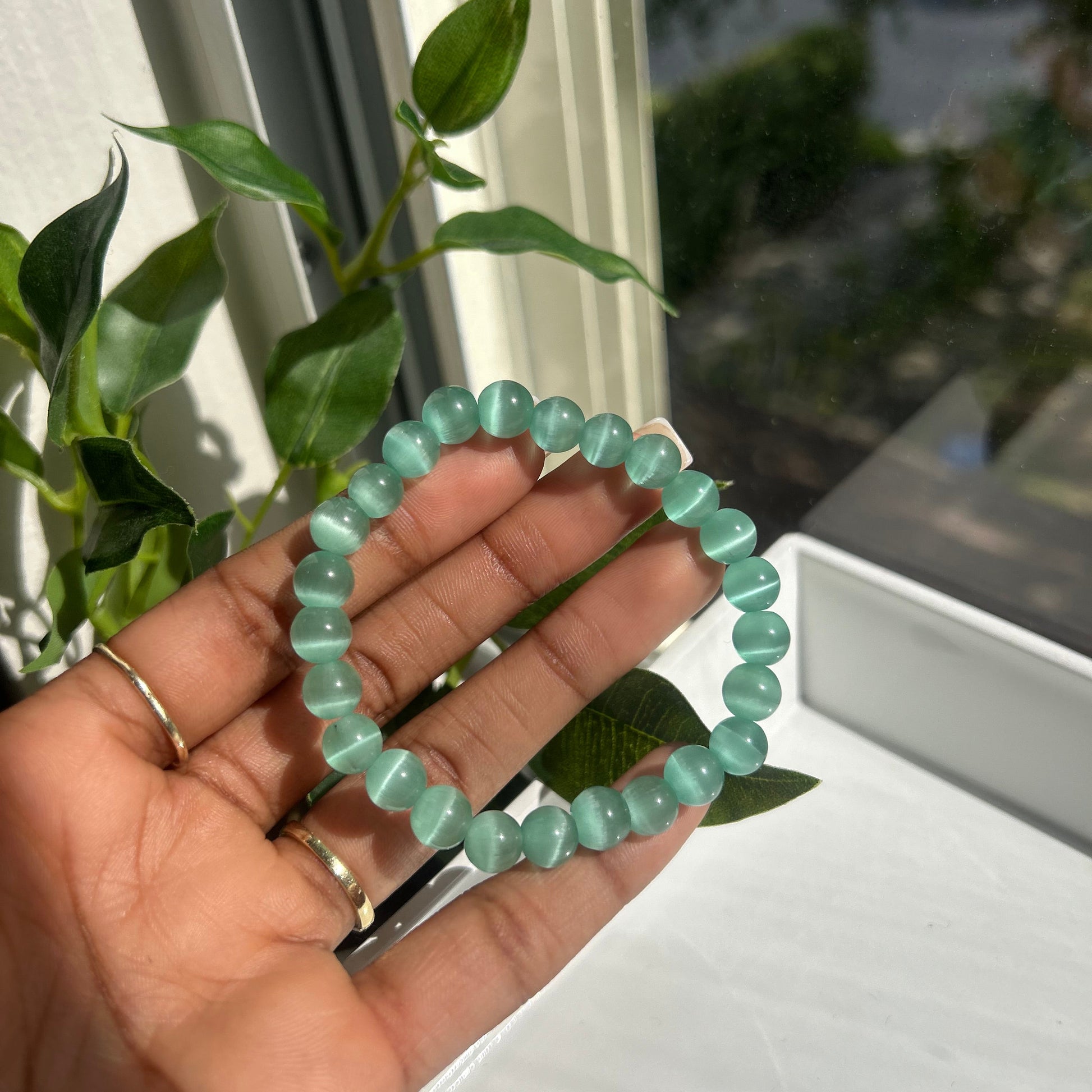A beaded bracelet made of light mint green translucent oval beads, held in a person's hand near a window with green leaves in the foreground.