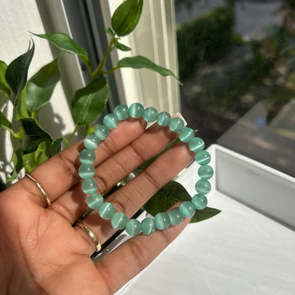 A beaded bracelet made of light mint green translucent oval beads, held in a person's hand near a window with green leaves in the foreground.
