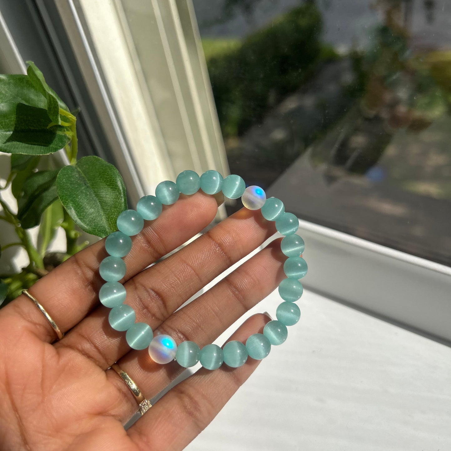 A person's hand holding a beaded stretch bracelet made of light blue-green oval beads with iridescent opal-like flashes, set against a window background.