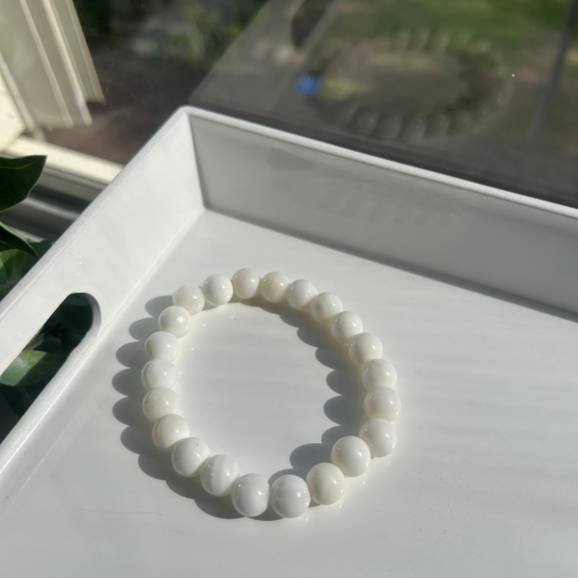 A white beaded bracelet made of smooth, rounded pearls or shell-like beads, displayed on a white tray in bright natural light.