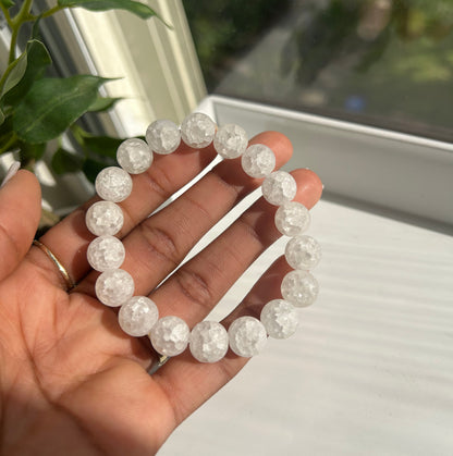 A beaded bracelet made of clear, faceted round beads held in a person's hand, with a window and green plant leaves in the background.