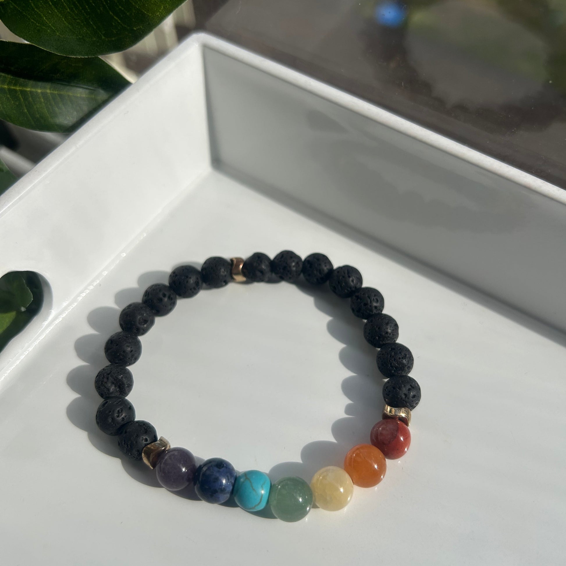A beaded stretch bracelet featuring dark navy blue stone beads and a segment of rainbow-colored gemstone beads, displayed on a white tray with green leaves in the foreground.