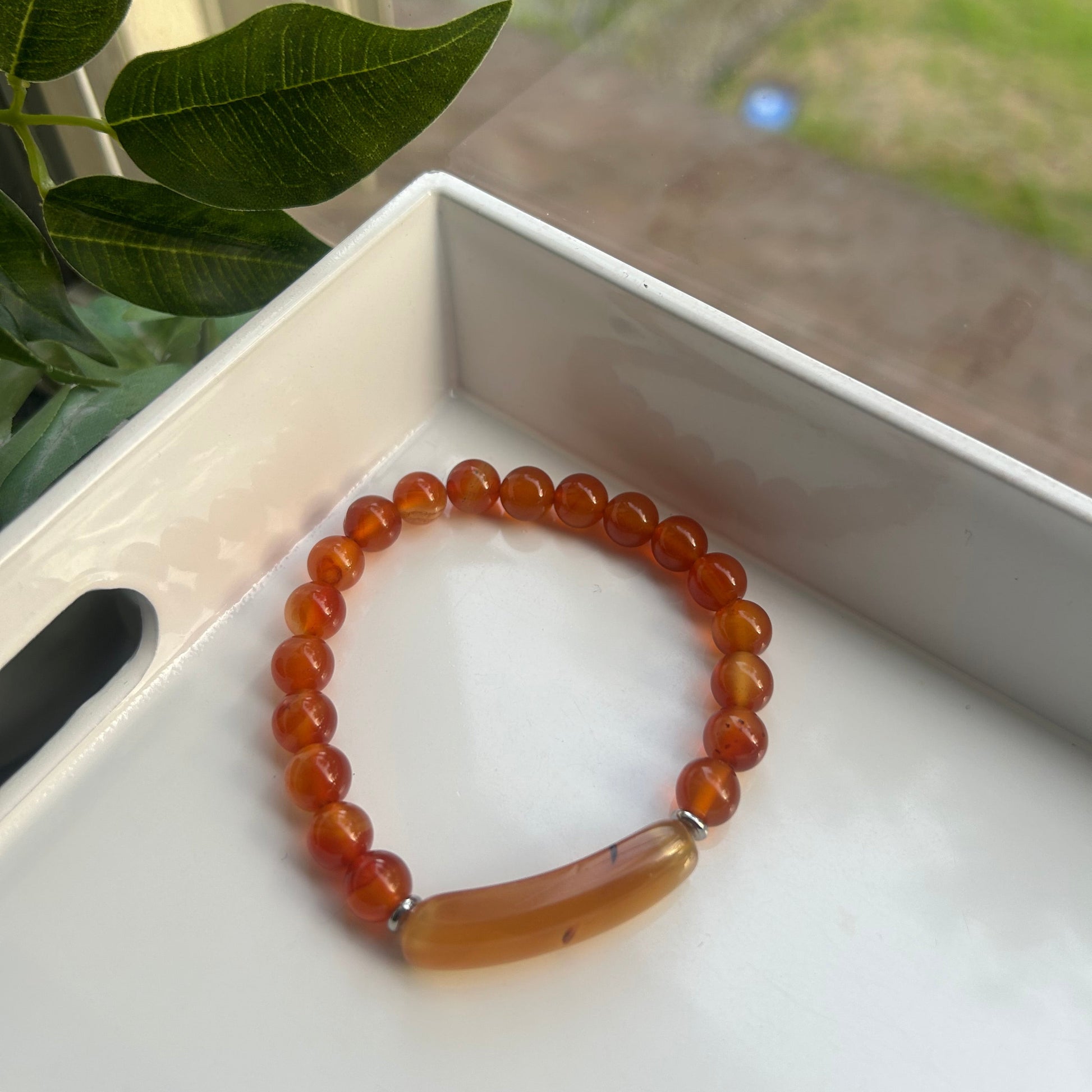A beaded stretch bracelet made of warm orange-brown translucent beads with a large polished oval stone end, resting inside a white tray near green leaves.