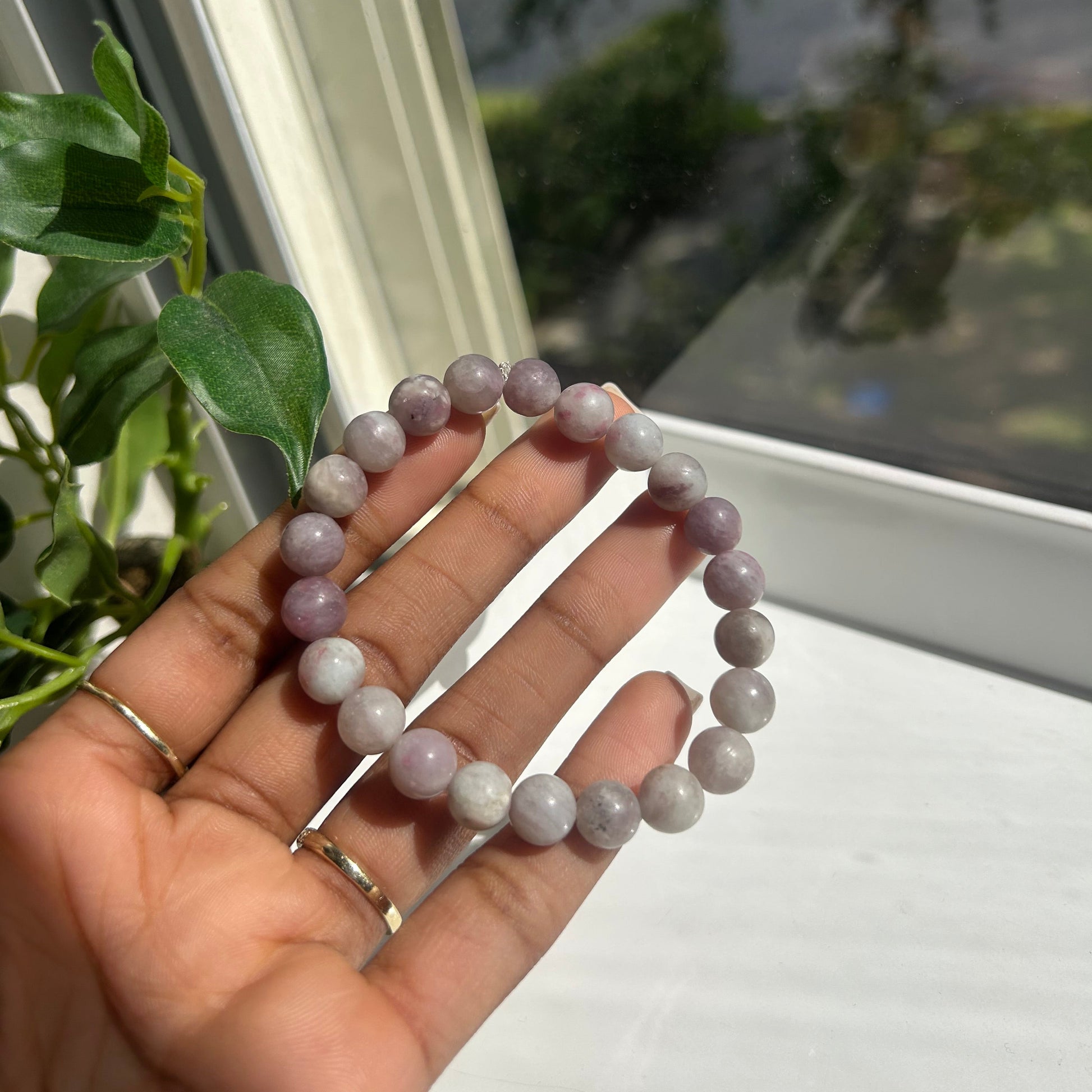 A beaded stretch bracelet made of round lavender-gray beads, held in a person's hand near a window with green leaves in the foreground.