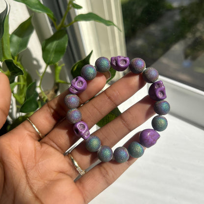 A handmade beaded bracelet with purple skull and rose motifs, held in a person's hand near a window with green leaves in the background.