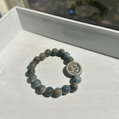 A beaded stretch bracelet made of round gray and blue-green beads with a central round silver-toned charm featuring an Om symbol, displayed on a white surface.