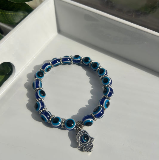 A blue and white evil eye beaded bracelet with a silver hamsa hand charm, displayed on a white tray with green leaves in the foreground.