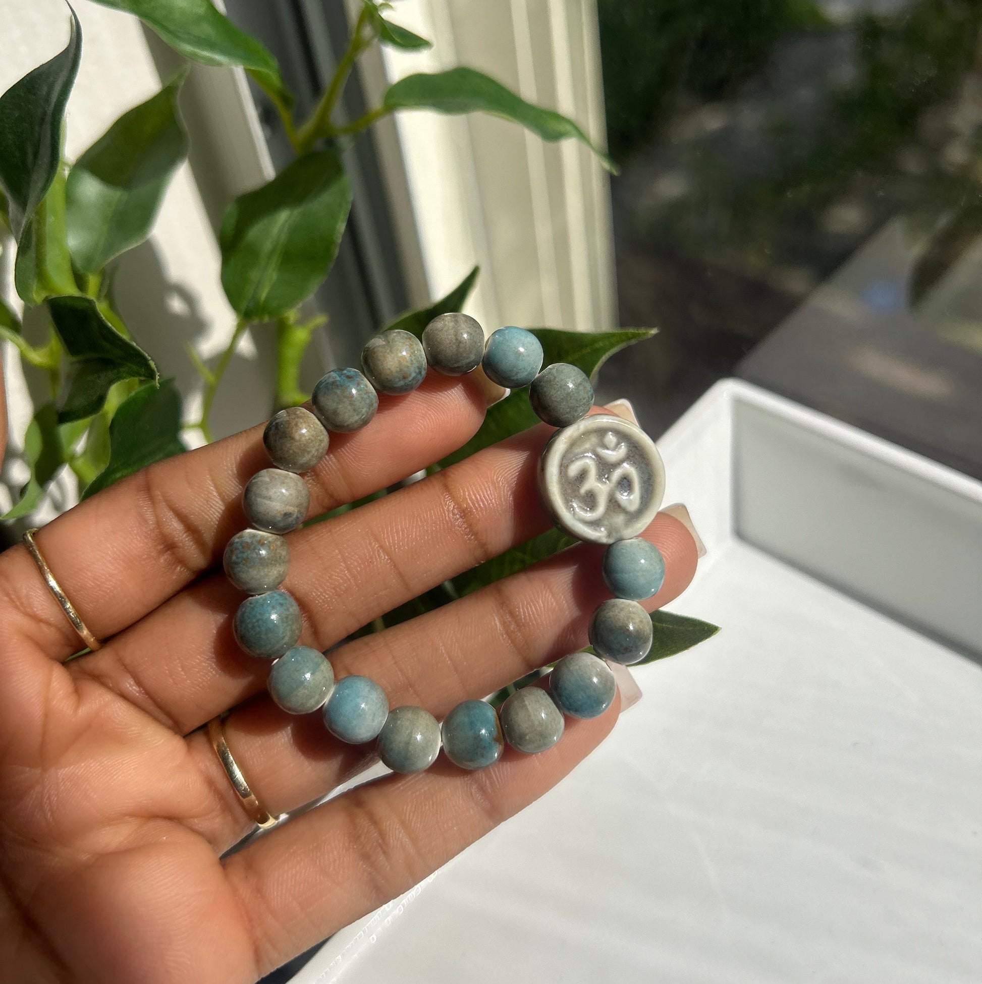 A beaded bracelet with light blue and gray marbled beads, featuring a white Om symbol charm, held in a person's hand with green leaves and a window in the background.