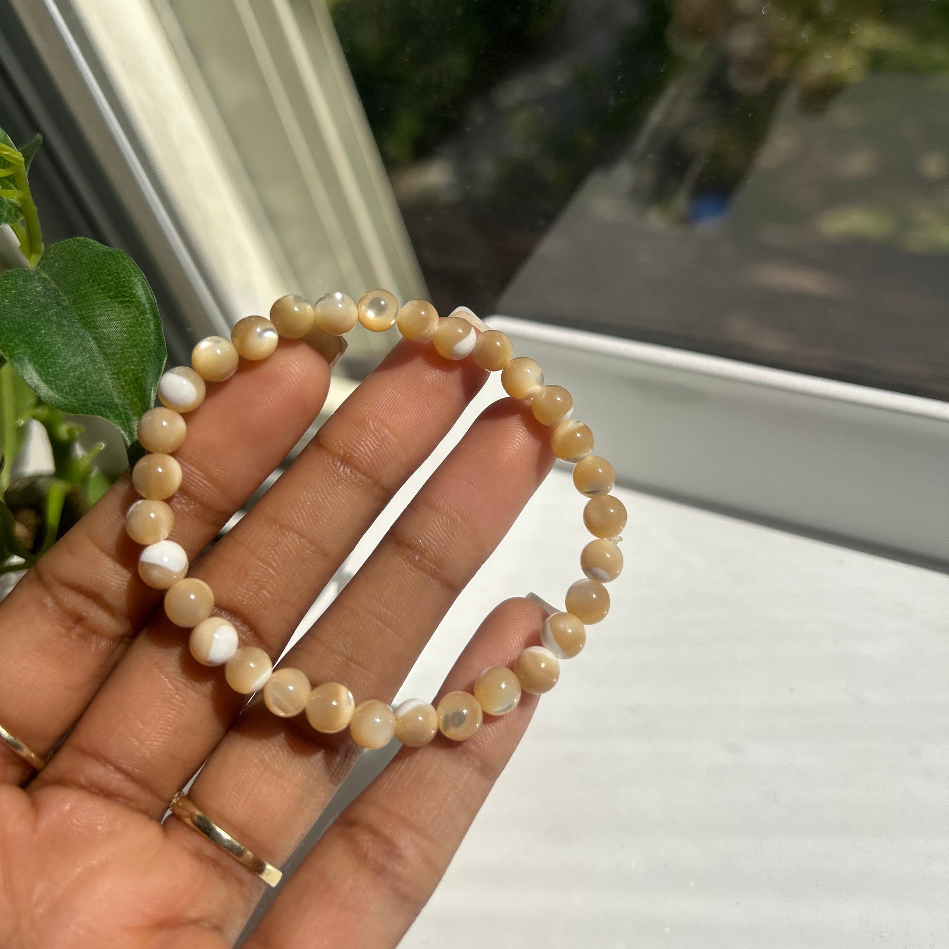 A person's hand holding a beaded stretch bracelet made of small, round, light tan and cream-colored beads with a few white accents, positioned in front of a window.